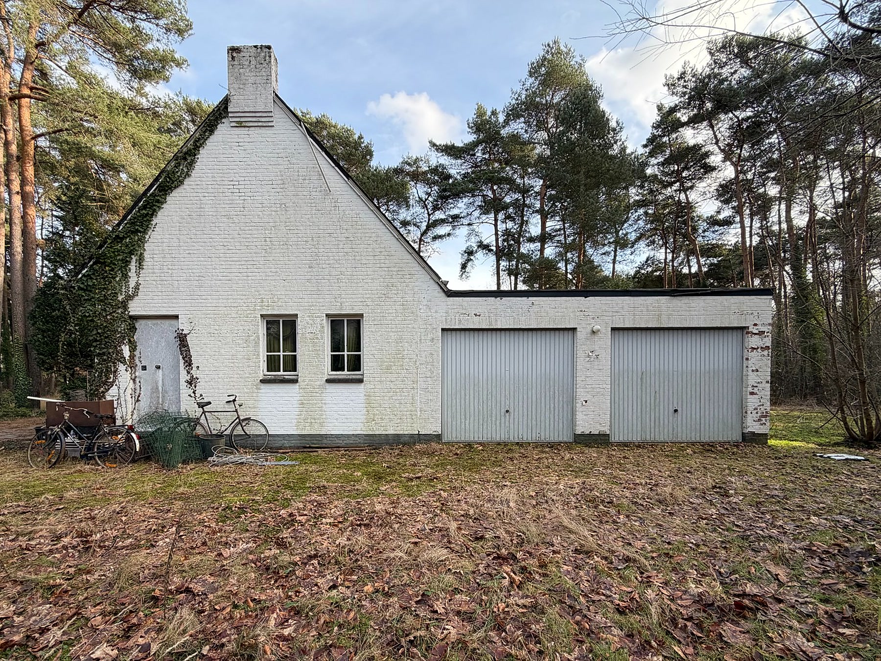 Wit bijgebouw met twee garagepoorten, omgeven door bos